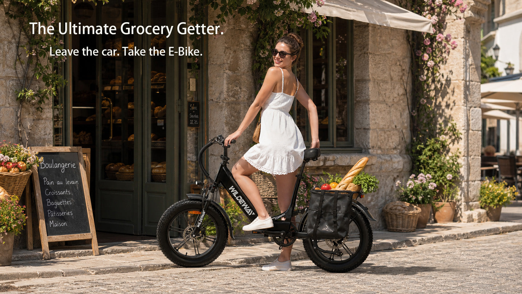 A black WILDEWAY Torpedo step-through e-bike parked outside a market, with its rear rack loaded with pannier bags full of fresh groceries and a baguette.